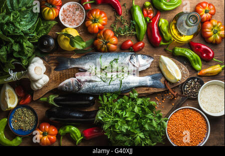 Rohe ungekochte Wolfsbarsch Fisch mit Gemüse, Getreide, Kräuter und Gewürze auf Schneiden an Bord über rustikalen hölzernen Hintergrund, Ansicht von oben Stockfoto