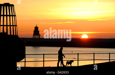 Ein Mann geht mit einem Hund, während die Sonne am Fish Quay nahe der Mündung des Flusses Tyne, North Shields, aufgeht. Stockfoto