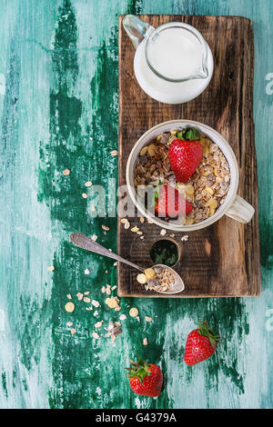Zutaten für gesundes Frühstück. Krug mit Milch, Schüssel Müsli und Erdbeeren auf Holz hacken Board Teelöffel über weiß und g Stockfoto