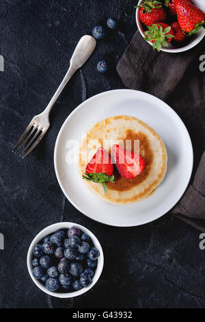 Pfannkuchen mit Erdbeeren und Heidelbeeren in weißer Keramik Teller und Schalen mit frischen Beeren auf Textile Serviette über schwarz tex Stockfoto