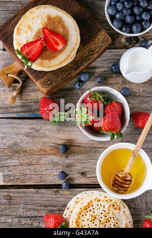 Pfannkuchen mit Erdbeeren und Heidelbeeren, Schalen mit Honig und Krug Milch über alten hölzernen Hintergrund. Frühstück Konzept Hinterg Stockfoto