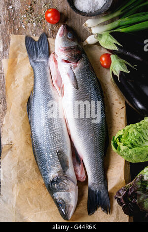 Zwei rohen ungekochten Seebass Fisch auf Backpapier mit Meersalz, getrocknete Kräuter und Gemüse über alten hölzernen Hintergrund. Ansicht von oben Stockfoto