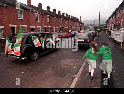 Ein mit Sinn-Fein-Wahlplakaten und -Fahnen bedecktes Taxi aus Belfast Black zieht zwei Schülerinnen an, wie Gerry Adams, der Sinn-Fein-Präsident, heute (Di) durch die terrassenförmigen Straßen von West Belfast fährt. Stockfoto
