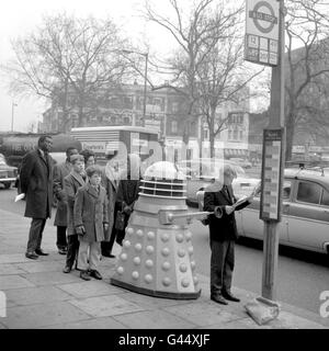 Ein junger anwartender Passagier liest seine Zeitung, ohne den Dalek aus der BBC-Fernsehsendung „Dr. Who“ zu kennen, in der Schlange hinter ihm. Stockfoto