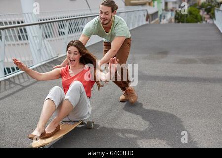 -MODELL VERÖFFENTLICHT. Junge Frau sitzt auf Skateboard mit Mann schieben. Stockfoto