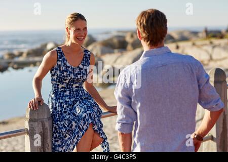 -MODELL VERÖFFENTLICHT. Frau sitzt am Geländer vom Strand lachen, Mann beobachtet. Stockfoto