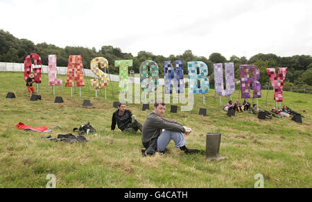 Nachtschwärmer sitzen auf dem Gras in der Nähe eines Hinweises für das Glastonbury Festival in Somerset. Stockfoto