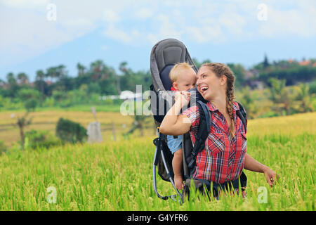 Naturspaziergang in grünen Terrasse Reisfeld. Glückliche Mutter halten wenig Reisende im Rucksack. Baby Ride auf Frau zurück. Stockfoto