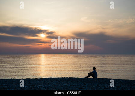 Junger Mann sitzt auf dem Stein Strand am Meer Sonnenuntergang Hintergrund Stockfoto