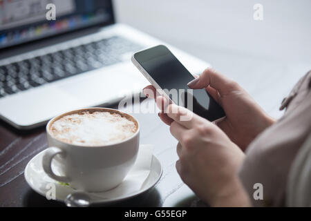 Weibliche Hände halten Smartphone im Coffee-Shop mit Laptop auf dem Hintergrund. Frau sitzt mit Tasse Kaffee Stockfoto