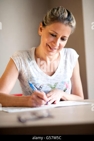 Einzelne schöne Reife Frau mit hübschen Lächeln und gebundene Haare zurück schreiben von Notizen oder Ausfüllen eines Formulars mit Stift am Tisch im Innenbereich
