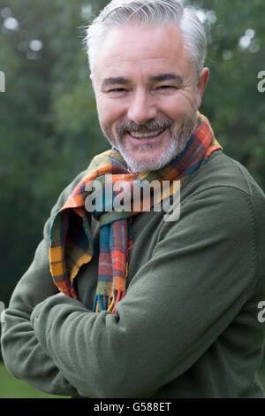 Porträt eines Mannes mit seine Arme verschränkt, Lächeln für die Kamera. Er ist draußen und Bäume im Hintergrund zu sehen. Stockfoto