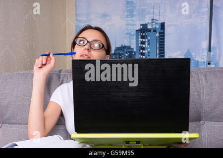 Verträumte junge Frau hat in Gedanken verloren, während auf einem Laptop arbeiten und ihr Geschäft von zu Hause mit einem Stift auf einem Sofa sitzt. Stockfoto