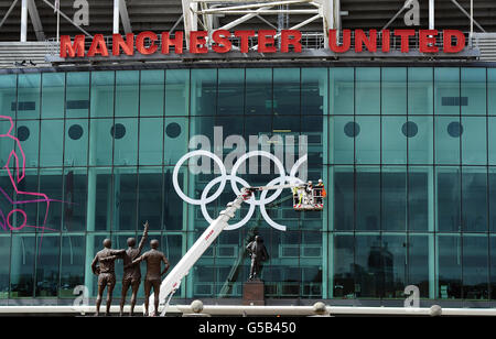 Riesige olympische Ringe sind hinter der Statue von Sir Matt Busby auf der Vorderseite des Old Trafford Football Ground, Heimat von Manchester United, in Manchester platziert. Stockfoto