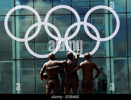 Riesige olympische Ringe befinden sich hinter der Statue der United Trinity auf der Vorderseite des Old Trafford Football Ground, dem Heimstadion von Manchester United, in Manchester. Stockfoto