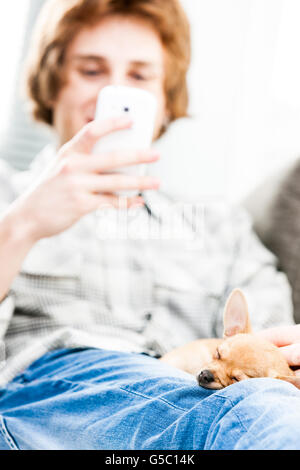 Niedliche kleine braune Chihuahua an den Beinen eines jungen Mannes mit einem Mobiltelefon entspannen auf dem Sofa, den Fokus auf den Hund schlafen Stockfoto