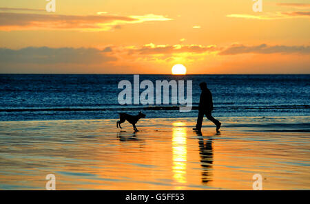 Ein Mann geht seinen Hund entlang Tynemouth Strand, North Tyneside, als die Sonne nach einer Woche von schlechtem Wetter aufgeht. Stockfoto