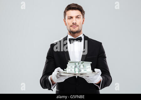Attraktive junge Butler in Smoking und Handschuhe halten Geld auf Tablett Stockfoto