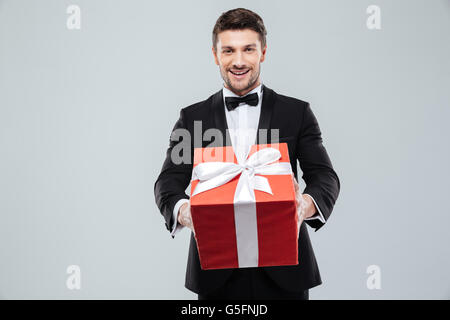 Fröhlich attraktive junge Mann im Smoking geben Ihnen rote Geschenkbox Stockfoto