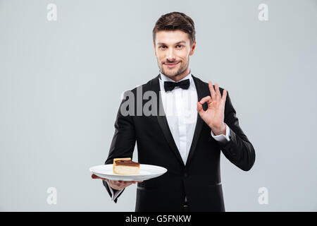 Hübscher junge Kellner im Smoking mit Fliege Halteplatte mit Kuchen und zeigt ok Sign. Stockfoto