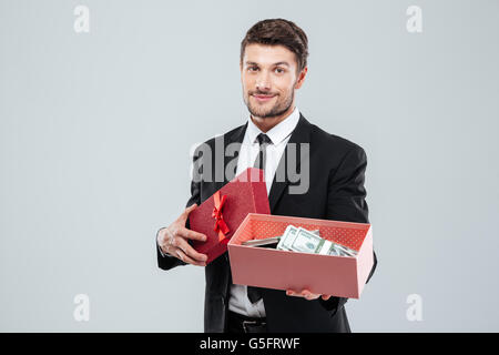 Lächelnde junge Geschäftsmann zeigt Geschenkbox voller Geld auf weißem Hintergrund Stockfoto