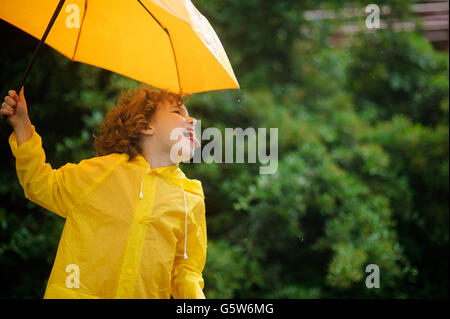 Fröhlicher Junge mit einem leuchtend gelben Regenschirm in Regenmantel. Laddie ragen Zunge und Fänge von Regentropfen. Er ist durch ein solches Spiel unterhalten. Stockfoto