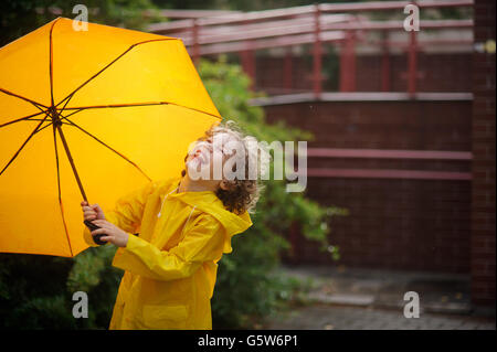 Der junge mit einem gelben Schirm freut sich, einen Regen. Das Kind ist in einem hellen gelben Regenmantel gekleidet. Er ist im Hof und lachen Stockfoto