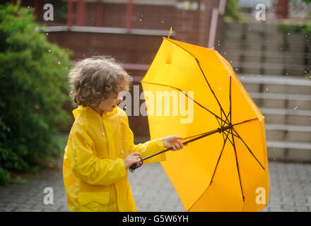Der junge in einem hellen gelben Regenmantel öffnet einen Regenschirm in den Regen. Das Kind ist auf dem Hof des Hauses. Ein Regentropfen fallen Stockfoto