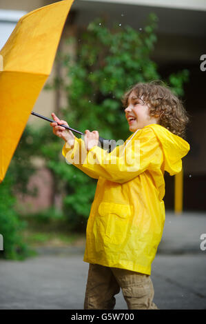 Laddie versucht 8-9 Jahre in einem hellen gelben Regenmantel, einen Regenschirm von Windflaws zu halten. Der junge hält einen Regenschirm mit beiden Händen. Stockfoto