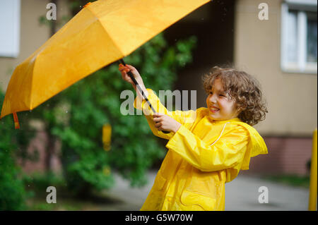 Der junge versucht 8-9 Jahre in einem hellen gelben Regenmantel, einen Regenschirm aus Wind Fehler halten. Er hält einen Regenschirm mit beiden Händen. Ch Stockfoto