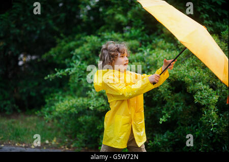 Lockige junge in einem hellen gelben Regenmantel mit einem Regenschirm gegen einen herrlichen grünen Busch. Stockfoto