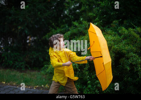 Binsen von starkem Wind ziehen Sie einen Regenschirm aus dem jungen Händen. Kerlchen gekleidet in einem hellen gelben Regenmantel stark hält eine Stockfoto