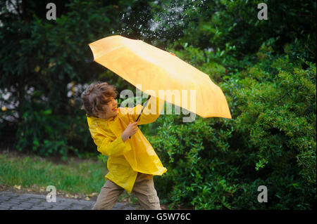Junge versucht 8-9 Jahre in einem hellen gelben Regenmantel, einen Regenschirm aus Wind Fehler halten. Er kleidete sich in einem hellen gelben Regenmantel. Lar Stockfoto