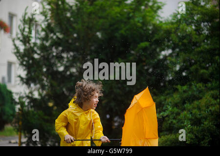 Laddie in einem hellen gelben Regenmantel und mit einem Regenschirm ist beständig gegen eilt der starke Wind. Kaum hält er einen Regenschirm. Eine faire ha Stockfoto