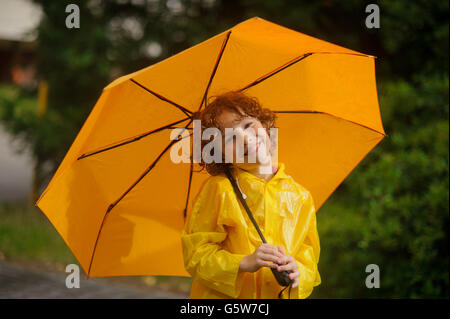 Der junge mit einem gelben Schirm. Er ist in einem hellen gelben Regenmantel gekleidet. Der junge hat ein schönes Gesicht und lockiges Haar. Stockfoto