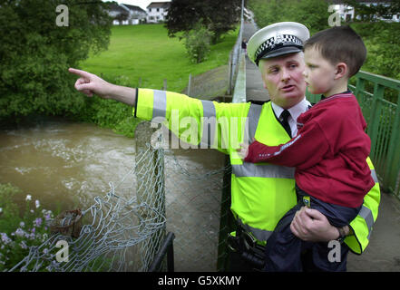 Der sechsjährige Andrew Davidson wird in den Armen von Sergeant Gordon McClure gehalten, der auf die Stelle zeigt, an der er ihn vom Black Devon River in Clackmannan in der Nähe von Stirling gezogen hat, nachdem der Junge einfiel. Stockfoto