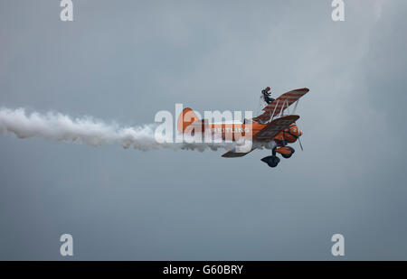 Orange Breitling Wingwalker Boeing Stearman Doppeldecker Kunstflug am Himmel mit einer Frau zu Fuß auf dem Flügel zu tun. Stockfoto