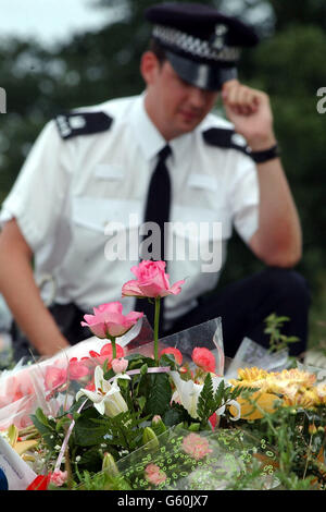 Ein Polizist schaut auf Blumen, die auf dem Gelände in der Nähe von RAF Lakenheath zurückgelassen wurden, wo am Samstag zwei Leichen gefunden wurden. Die Polizei hat enthüllt, dass sie „so sicher wie möglich sind“, dass es sich bei den beiden Leichen um die vermissten 10-jährigen Jessica Chapman und Holly Wells handelt. Stockfoto