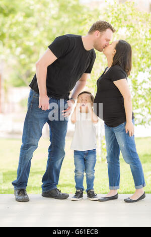 Junge Mischlinge Sohn versteckt Augen als schwangere Mama und Papa Kuss. Stockfoto