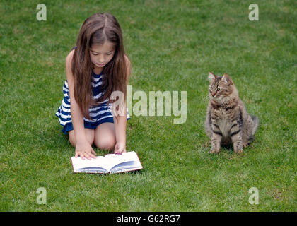 Kleines Mädchen mit Tabby Katze sitzt auf dem Rasen, Buch lesen, Stockfoto