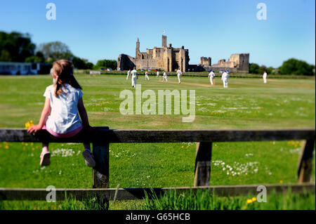 Ein Cricket-Spiel findet vor dem Hintergrund des verfallenen mittelalterlichen Gebäudes Warkworth Castle in Northumberland statt. Stockfoto