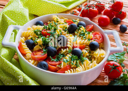 Nudeln (Fusilli) in weißen Pfanne auf hölzernen Hintergrund. Vegetarisch, mit Tomaten, schwarzen Oliven, Knoblauch und Petersilie gekocht. Stockfoto