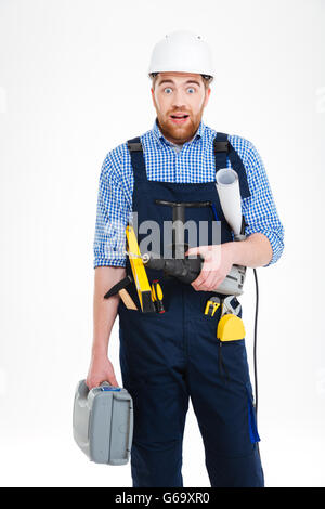 Erstaunt bärtigen jungen Baumeister in Helm mit Werkzeugkasten, Bohrmaschine und Baupläne Stockfoto