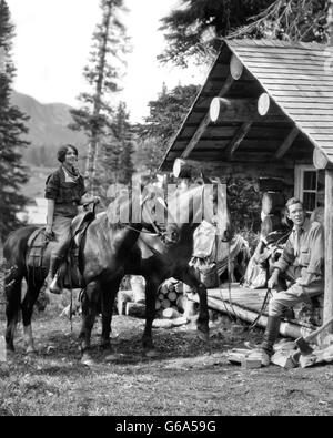 1930ER JAHREN PAAR VOR BLOCKHÜTTE FRAU AUF PFERD MANN SITZT AUF DER VERANDA, DIE ZÜGEL DER ZWEITEN PFERD ASSINIBOINE KANADA Stockfoto