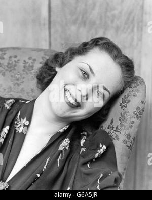 1930S 1940S PORTRAIT LÄCHELND BRÜNETTE FRAU GELEHNT ZURÜCK OHRENSESSEL BLICK IN DIE KAMERA SEITLICH Stockfoto