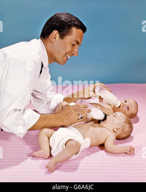 1960ER JAHREN VATER FÜTTERUNG ZWILLINGEN MIT FLASCHEN GLEICHZEITIG Stockfoto