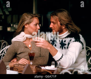 1970ER JAHREN ROMANTISCHES PAAR MANN FRAU MIT GETRÄNKE IM CAFÉ IM FREIEN SITZEN BEIDE TRAGEN PULLOVER MANN HAT LANGE HAARE UND BART Stockfoto