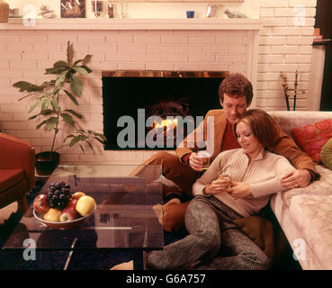 1970ER JAHREN ROMANTISCHES PAAR MANN UND FRAU, DIE GETRÄNKE KAMIN SITZEN AUF BODEN Stockfoto