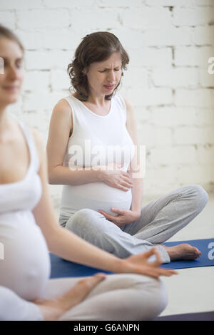 Schwangere Frau Gefühl Magenschmerzen bei Fitness oder Yoga Klasse drinnen. Junge schwangere Modell hält ihren Bauch, Unwohlsein Stockfoto