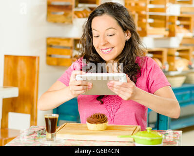 Hübsche Brünette Frau trägt rosa Hemd von Tabelle in einer Bäckerei, sitzen mit Handy zum Fotografieren der Brotscheibe mit Schokolade Stockfoto